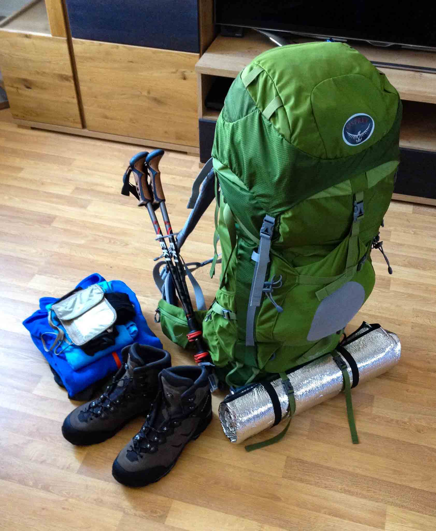 A green Osprey Aether 70l hiking backpack is packed with gear for a trip, including hiking boots, trekking poles, a sleeping pad, and clothing.